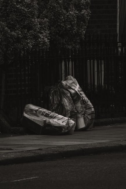 Two large black rubbish bags filled with waste are positioned on the pavement in front of a dark metal fence with vertical bars. The bags appear to be made of thick plastic, possibly black or dark gre