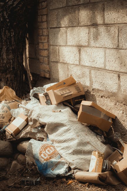 Two large black rubbish bags filled with waste are positioned on the pavement in front of a dark metal fence with vertical bars. The bags appear to be made of thick plastic, possibly black or dark gre